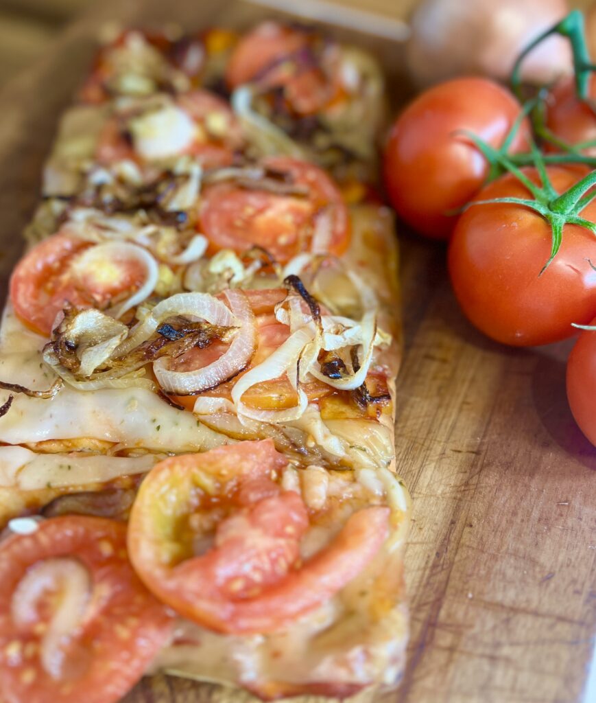 Flatbread pizza with tomato and caramelized onions on a cutting board next to fresh tomatoes on the vine.